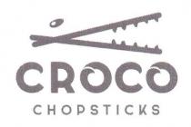 Logo raffigurante un chicco di riso e due bacchette con denti che simula il morso di un coccodrillo. Posto al Logo raffigurante un chicco di riso e due bacchette con denti che simula morso di un coccodrillo. Posto al di sotto del logo vengono riportate due righe contenenti un testo. Al primo rigo il testo CROCO ed al secondo rigo il testo CHOPSTICKS . Logo raffigurante un chicco di riso e due bacchette con denti che simula il morso di un coccodrillo. Posto al
